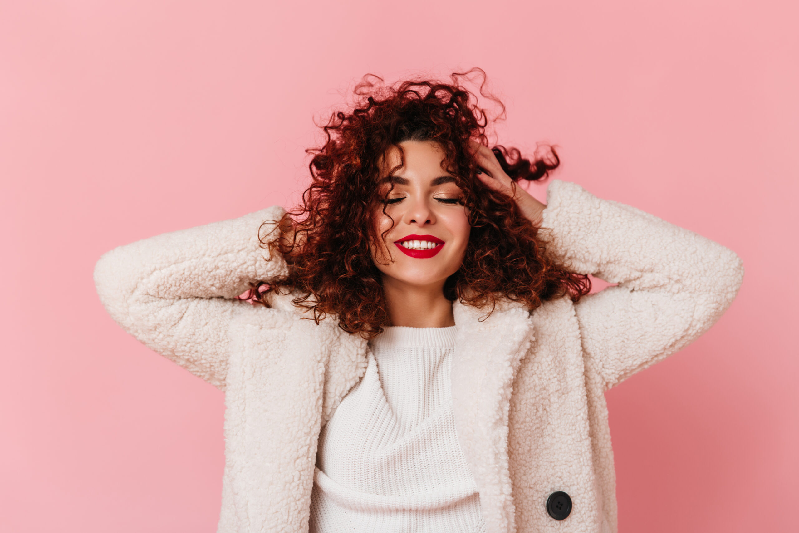 Portrait of charming lady with red lipstick and snow white smile dressed in bright eco coat and touching her curly hair on pink background.