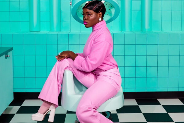 A picture by The Doux. Black woman elegantly sitting in a chair at a hair salon. She is wearing a bright pink suit, the hair salon has bringt aqua blue tiles on the walls and a black and white checkerd flooring.